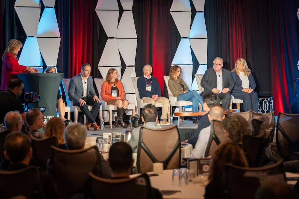 'n Paneel van sewe mense sit op die verhoog voor 'n geometriese agtergrond met rooi en blou beligting, teenoor 'n gehoor terwyl 'n vrou op 'n podium aan die linkerkant staan. Die vertrek is gevul met sittende deelnemers.