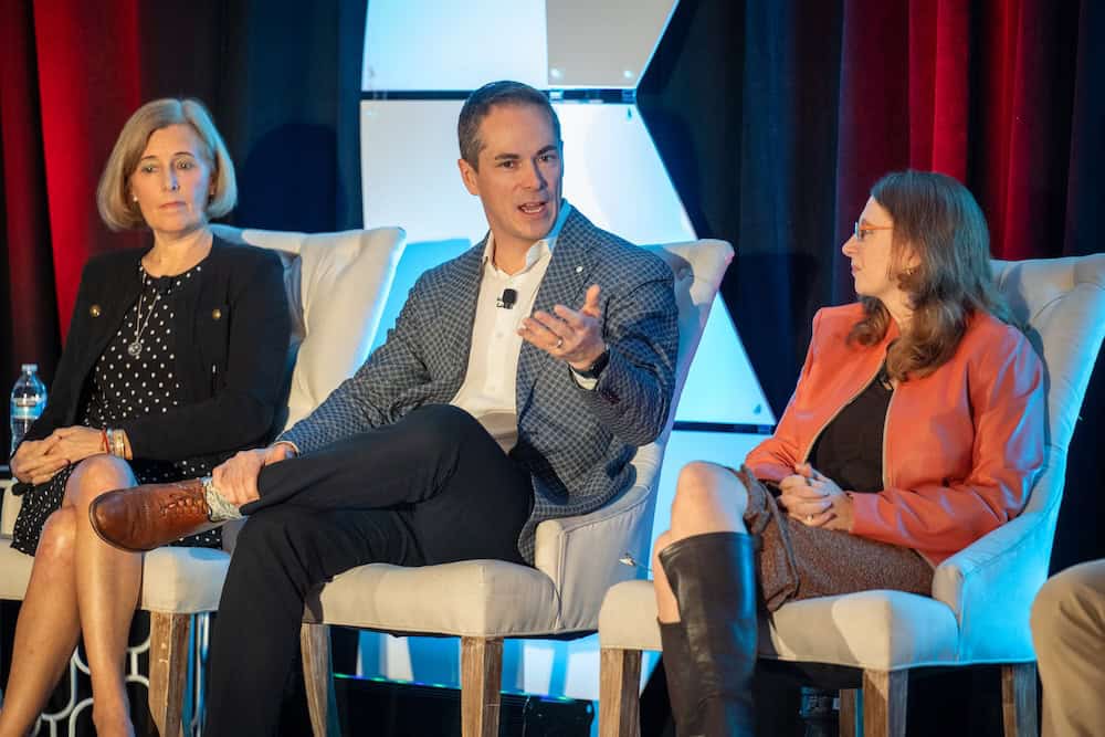 Drie mense sit op die verhoog in wit stoele, besig met 'n bespreking. Die man in die middel maak 'n gebaar terwyl hy praat, terwyl die twee vroue aan weerskante aandagtig luister. 'n Rooi en blou gordynagtergrond is agter hulle.