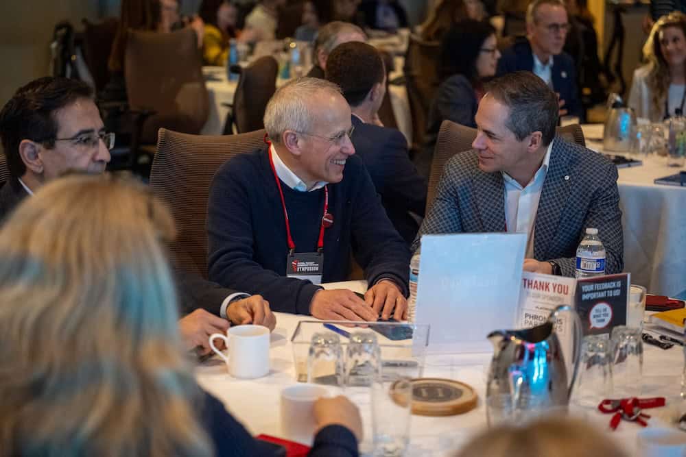 'n Groep mense sit om 'n ronde tafel by 'n konferensie of vergadering, besig met gesprekke en glimlaggend. Koffiebekers, glase en papiere is op die tafel, met ander wat in die agtergrond gesels.