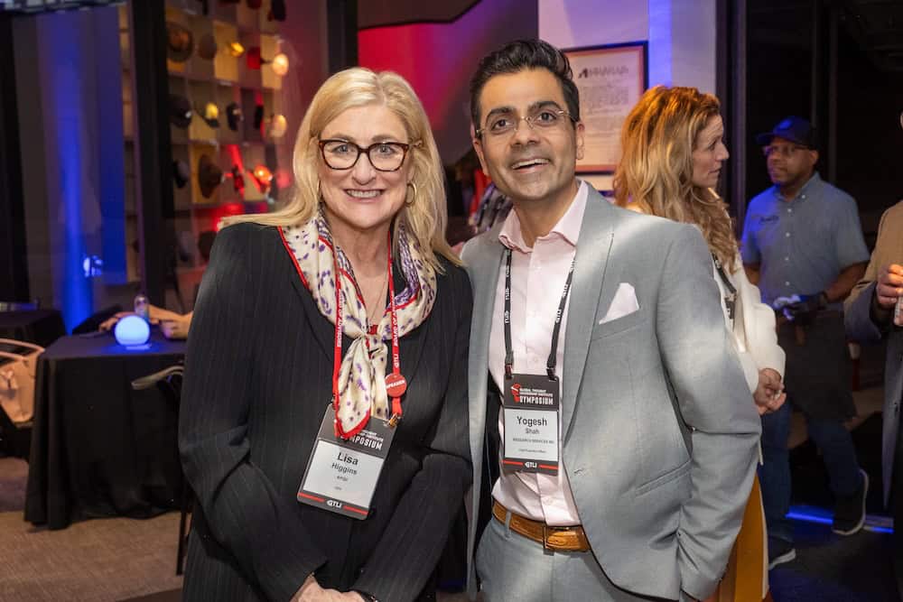 Twee mense met konferensiekentekens staan saam en glimlag vir die kamera in 'n lewendige binnenshuise geleentheidsomgewing, met ander deelnemers en kleurvolle ligte in die agtergrond.