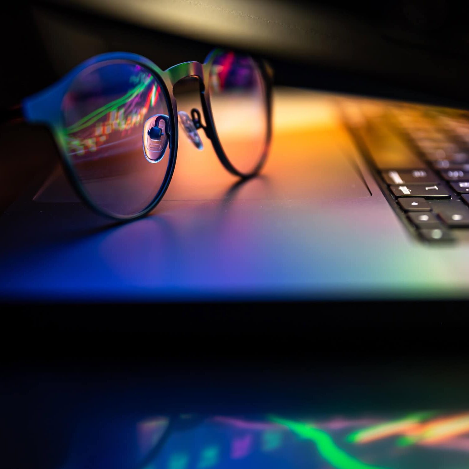 'n Nabyskoot van 'n bril wat op 'n skootrekenaar se sleutelbord rus, met kleurvolle ligrefleksies op beide die skootrekenaar se oppervlak en die bril, wat 'n lewendige, abstrakte effek skep.
