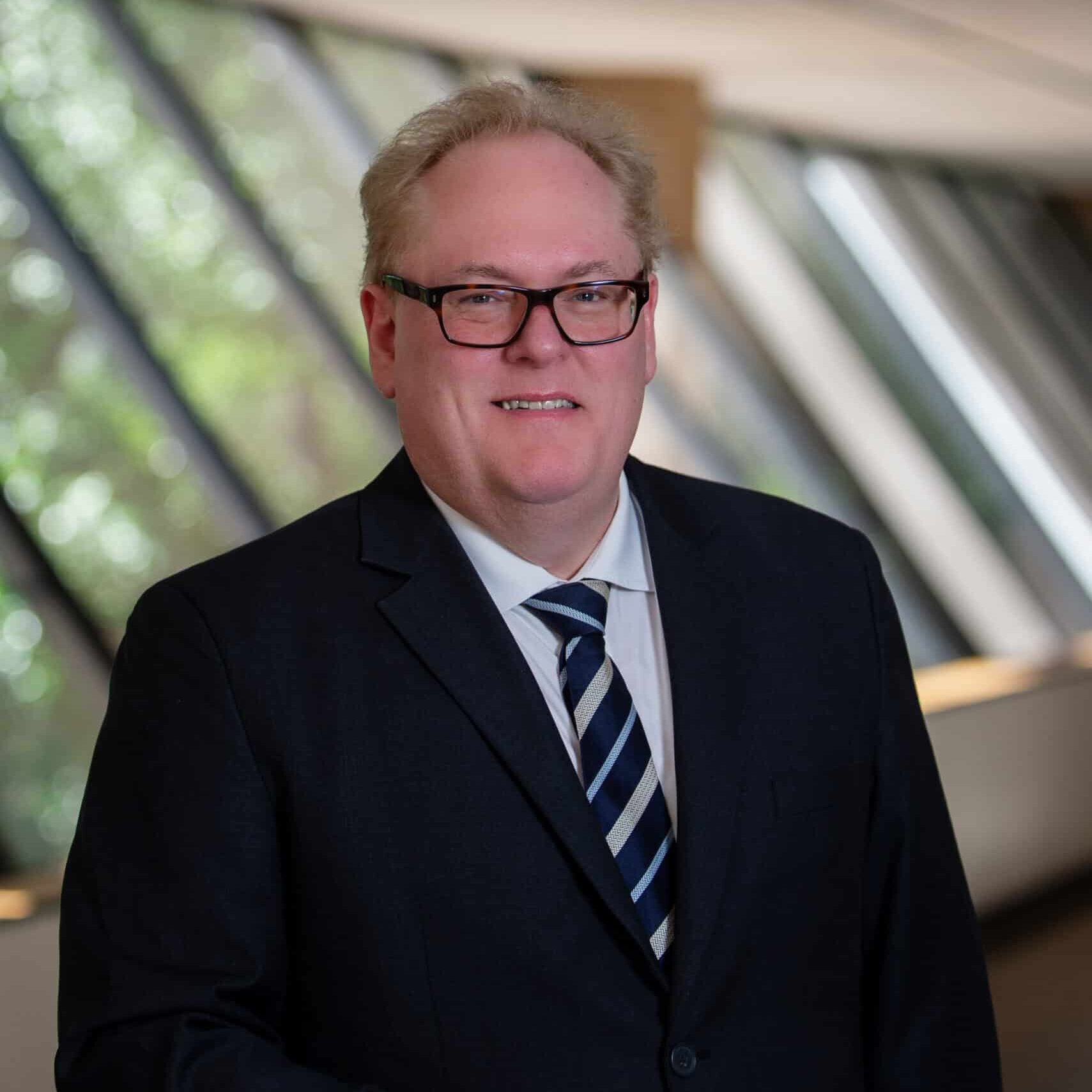 'n Man met ligte hare en 'n bril in 'n donker pak, wit hemp en gestreepte das staan binnenshuis langs 'n reling, met groot vensters en groenigheid sigbaar in die agtergrond.