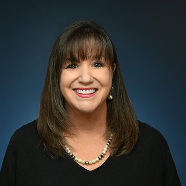 'n Vrou met reguit bruin hare en kuif glimlag vir die kamera. Sy dra 'n swart top, 'n pêrelketting, pêreloerbelle en het 'n donkerblou agtergrond agter haar.